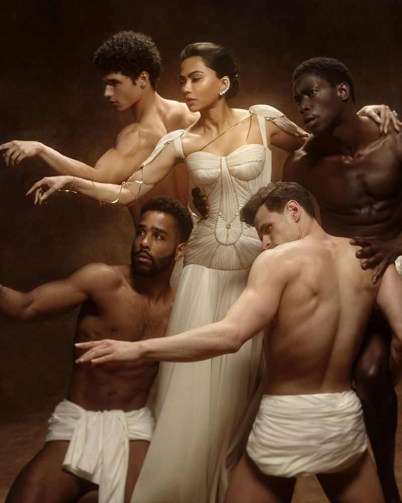Mona Patel in an ivory corseted gown surrounded by four shirtless male models posing in draped white cloths against a warm backdrop.