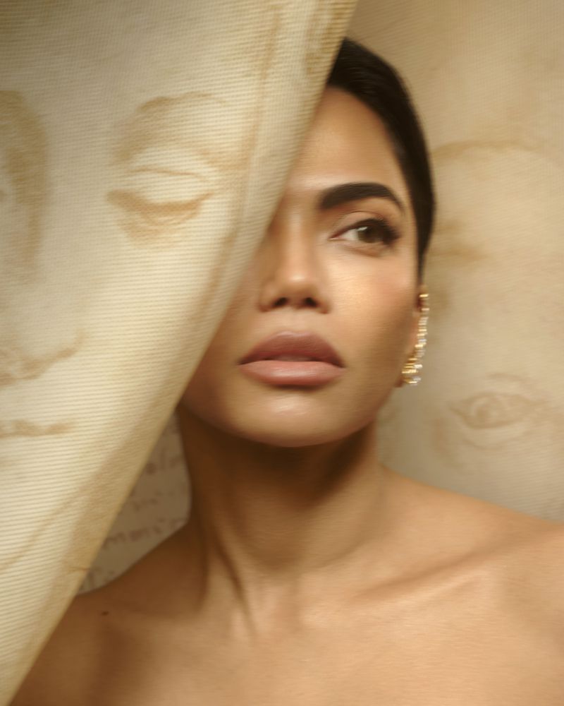 Close-up beauty portrait of Mona Patel peering behind an illustrated textured canvas with warm lighting.