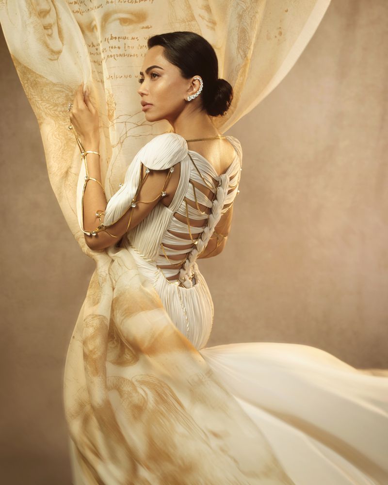 Mona Patel posing in a white pleated couture gown with an open strappy back while holding a printed Renaissance-style draped veil.
