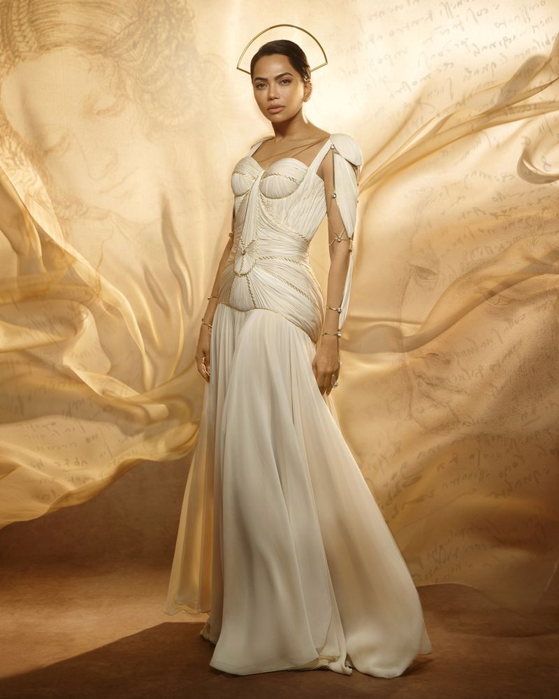 Mona Patel wearing a draped cream gown with a gold halo headpiece against a warm Renaissance sketch backdrop.