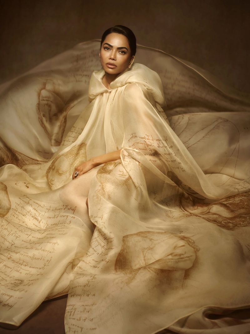 Mona Patel draped in an expansive cream sheer hooded cape printed with Renaissance sketches and manuscript script.