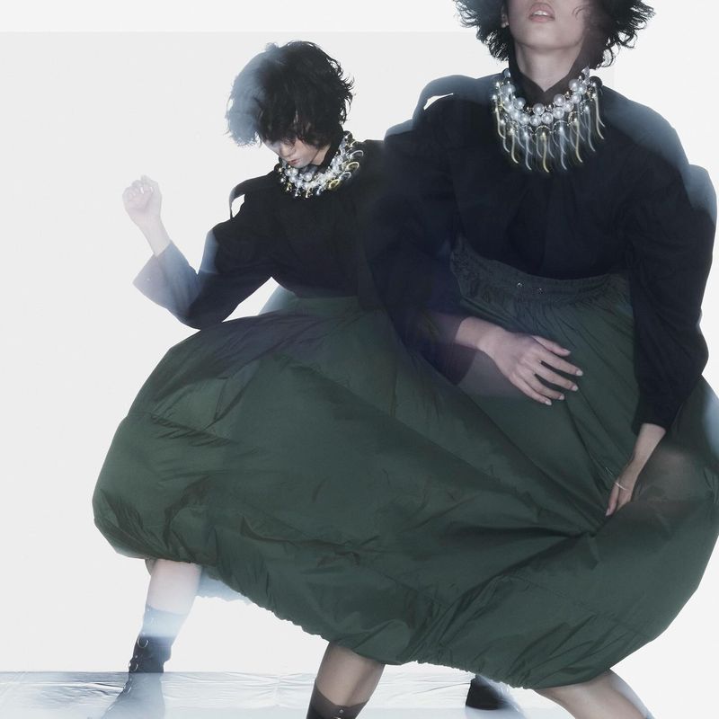Model dancing in a voluminous dark green balloon skirt, black blouse, and statement beaded necklace against a white background.