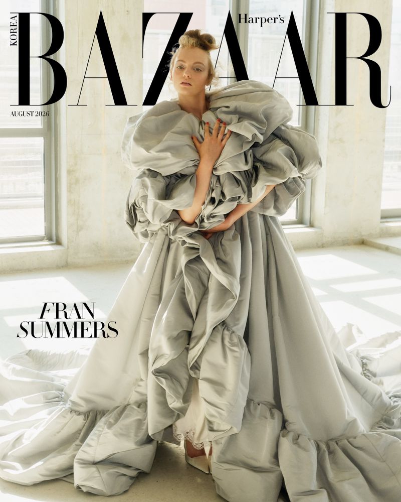 Model Fran Summers in a voluminous ruffled grey gown standing in a bright minimalist interior with large windows.