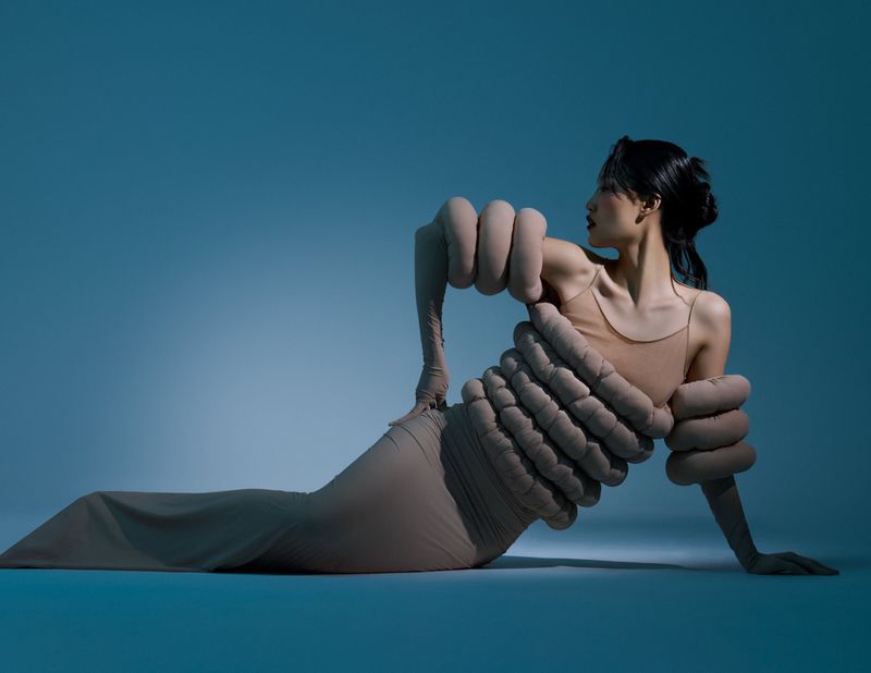A slender Asian woman in a beige dress poses creatively on a teal backdrop, with intriguing oversized fabric hand accents around her body.