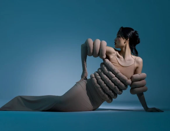 A slender Asian woman in a beige dress poses creatively on a teal backdrop, with intriguing oversized fabric hand accents around her body.