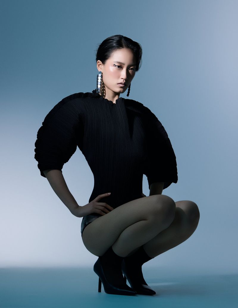 An Asian woman squats gracefully in a black outfit with stylish accessories, captured in a studio setting with soft, diffused lighting.
