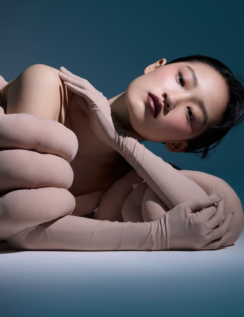 Elegant Asian woman in nude bodysuit and gloves poses gracefully against a teal backdrop in a beauty and fashion editorial shot.