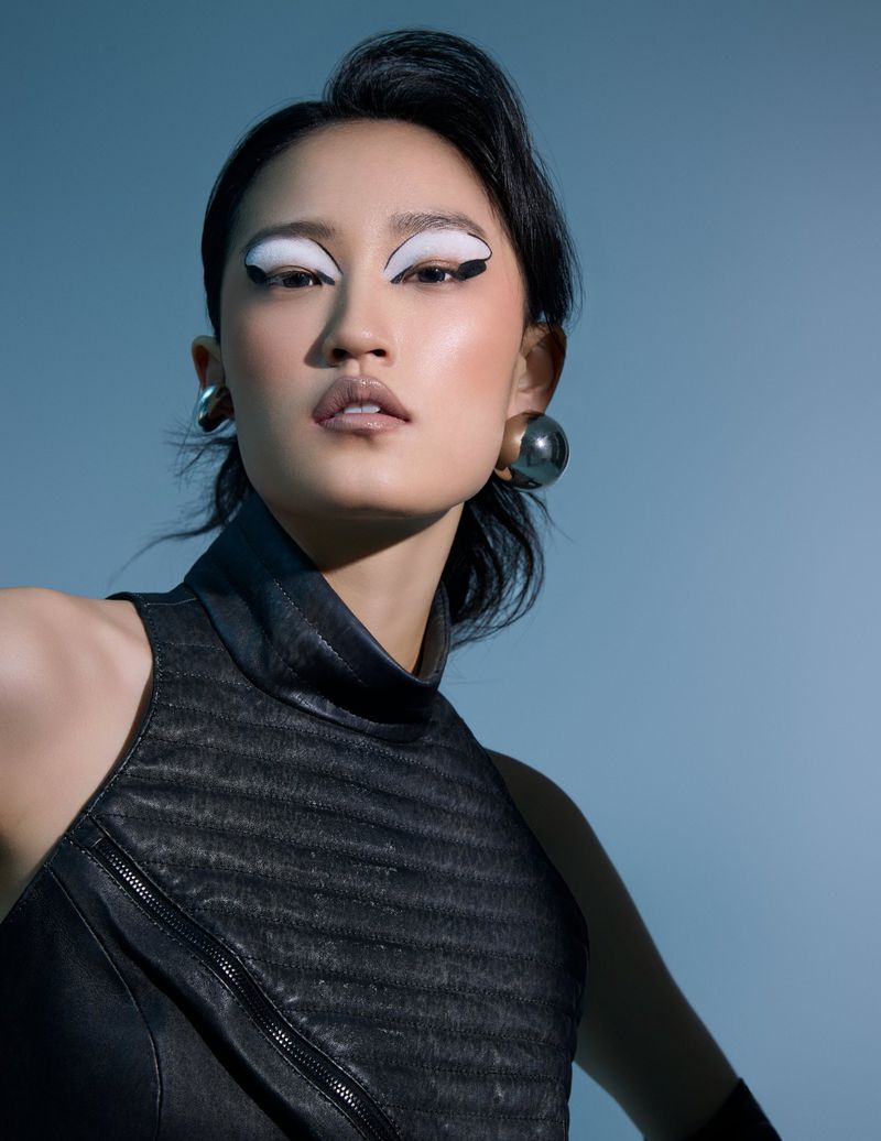 Asian woman in a fashion shot with black dress, bold white eye makeup, and large earrings, posing in front of a light blue background.