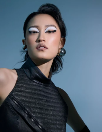 Asian woman in a fashion shot with black dress, bold white eye makeup, and large earrings, posing in front of a light blue background.