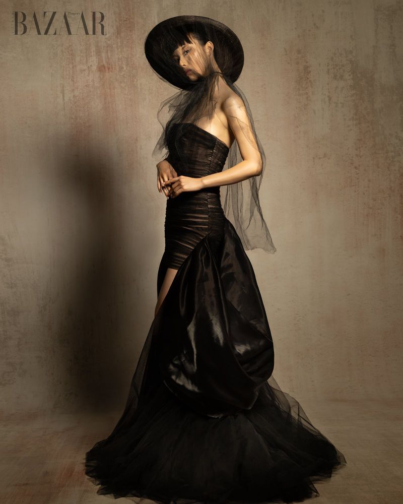 Model Bao Ziyi wearing a voluminous black strapless gown and veiled wide-brim hat against a textured backdrop for Harper's Bazaar.