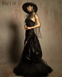 Model Bao Ziyi wearing a voluminous black strapless gown and veiled wide-brim hat against a textured backdrop for Harper's Bazaar.