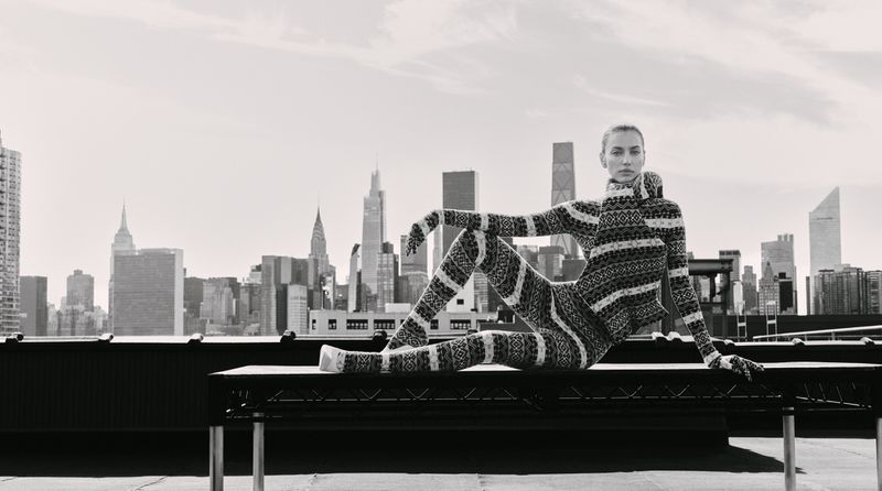 Irina Shayk wearing a patterned knit bodysuit posing on a rooftop bench against the Manhattan skyline.