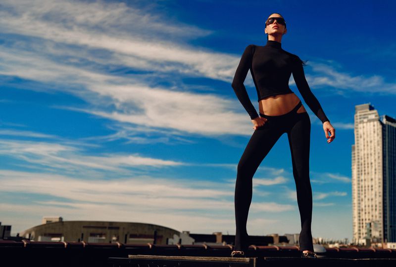 Model Irina Shayk wearing a black crop top, sunglasses, and black cut-out leggings poses against a bright blue sky on a city rooftop.