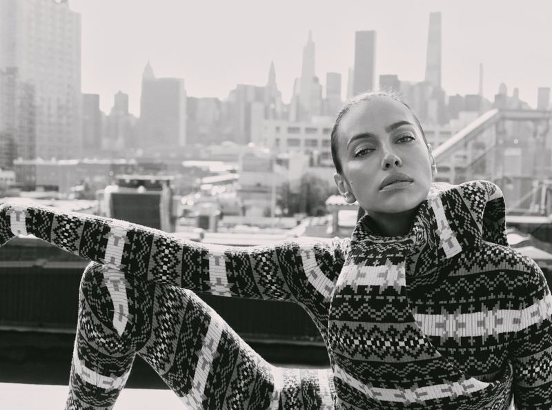 Model Irina Shayk wearing a patterned knit outfit sitting on an urban rooftop with a city skyline background.