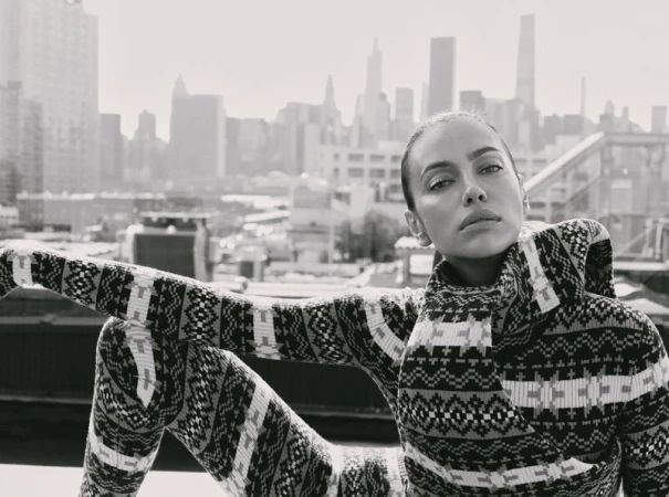 Model Irina Shayk wearing a patterned knit outfit sitting on an urban rooftop with a city skyline background.