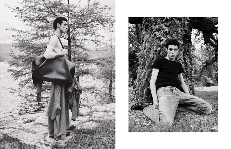 Black and white fashion shot of an adult male outdoors, posing with a large bag and wearing a long coat. Man leaning against a tree.