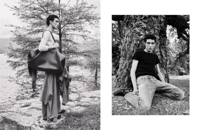 Black and white fashion shot of an adult male outdoors, posing with a large bag and wearing a long coat. Man leaning against a tree.