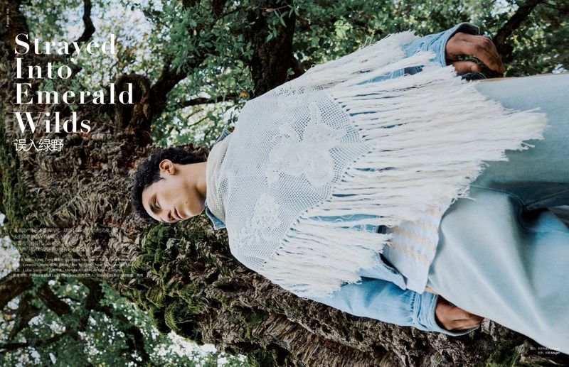 Outdoor fashion shot featuring a man wearing a denim jacket and fringed shawl in a lush, green forest setting with dappled light.