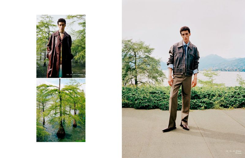 Man in fashionable outdoor editorial photoshoot featuring a brown jacket and trousers near the water with lakeside trees and natural light.