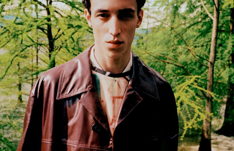 Close-up portrait of an adult male wearing a maroon leather jacket outdoors with a forest background, capturing a stylish and natural aesthetic.