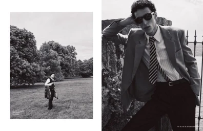 Black and white editorial photo of a man in a blazer and sunglasses leaning against a stone wall outdoors.
