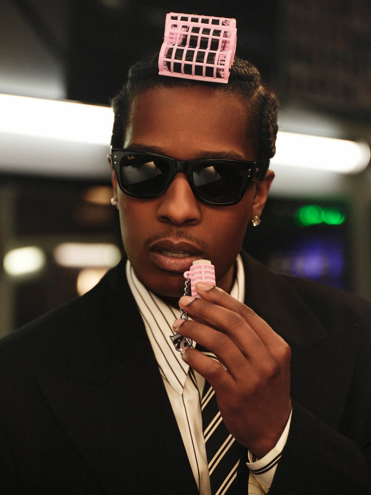 ASAP Rocky wearing black sunglasses and a black suit, holding a pink lip balm product with a pink hair roller on his head.
