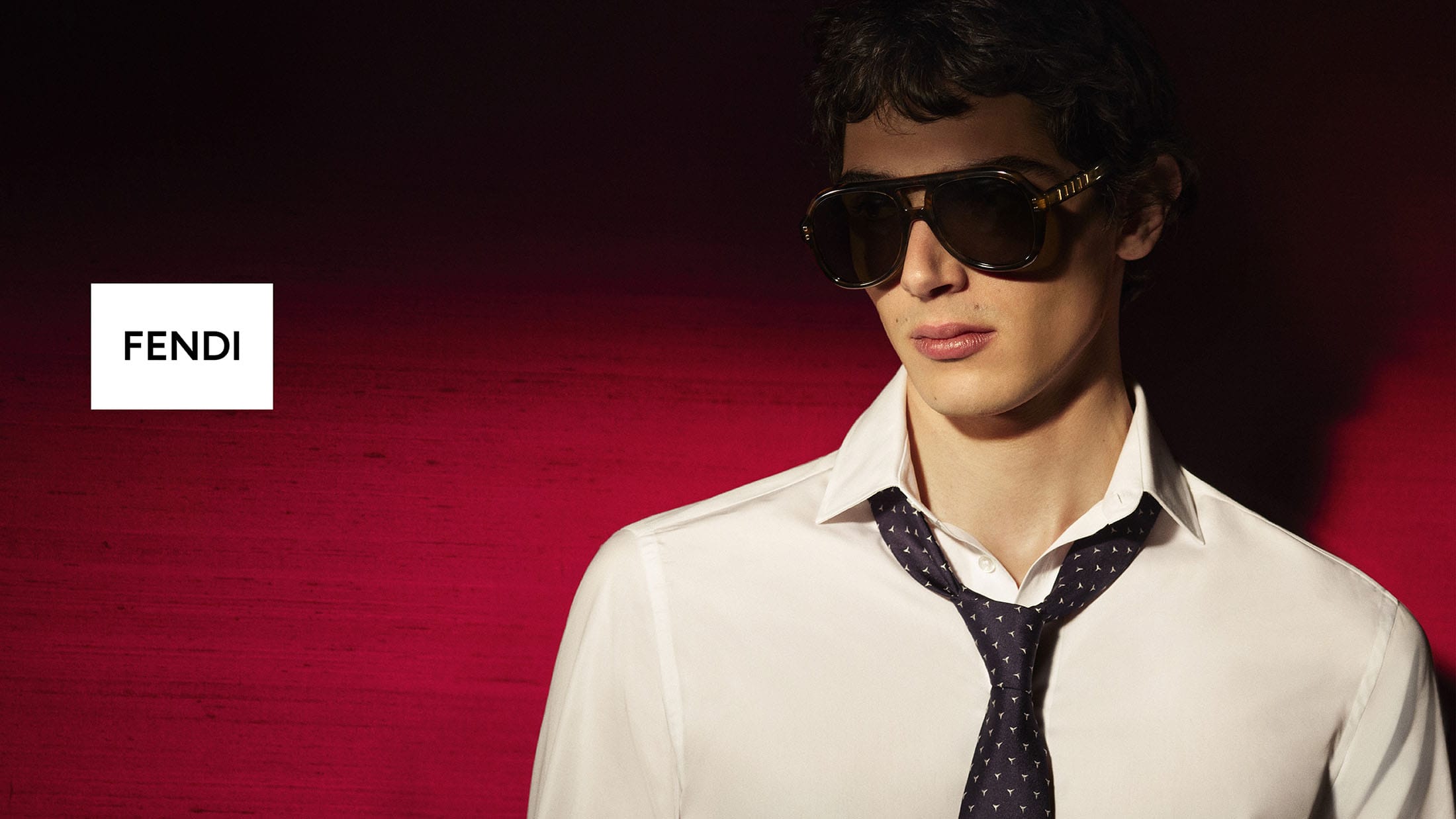 Model wearing dark oversized sunglasses, a white collared shirt, and a loosened necktie against a deep red backdrop with Fendi logo.
