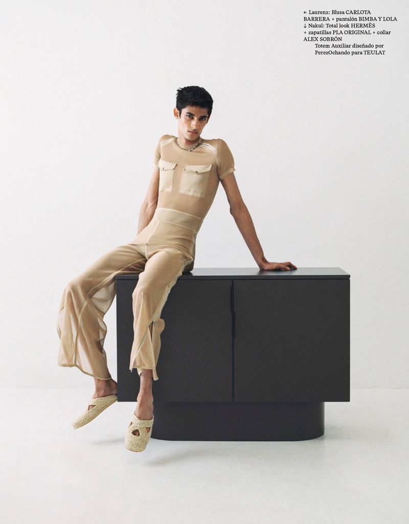 A stylish person sits on a black cabinet in a studio. The individual wears tan, see-through shirt, pants, and loafers, set against a white backdrop.