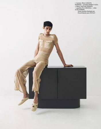 A stylish person sits on a black cabinet in a studio. The individual wears tan, see-through shirt, pants, and loafers, set against a white backdrop.