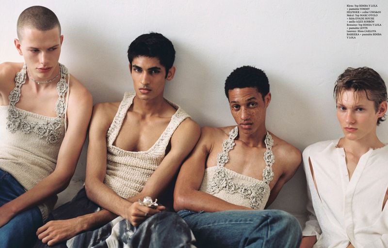 Four young adult men pose in an indoor fashion editorial. They wear crocheted tops and jeans, with a neutral background and soft lighting.