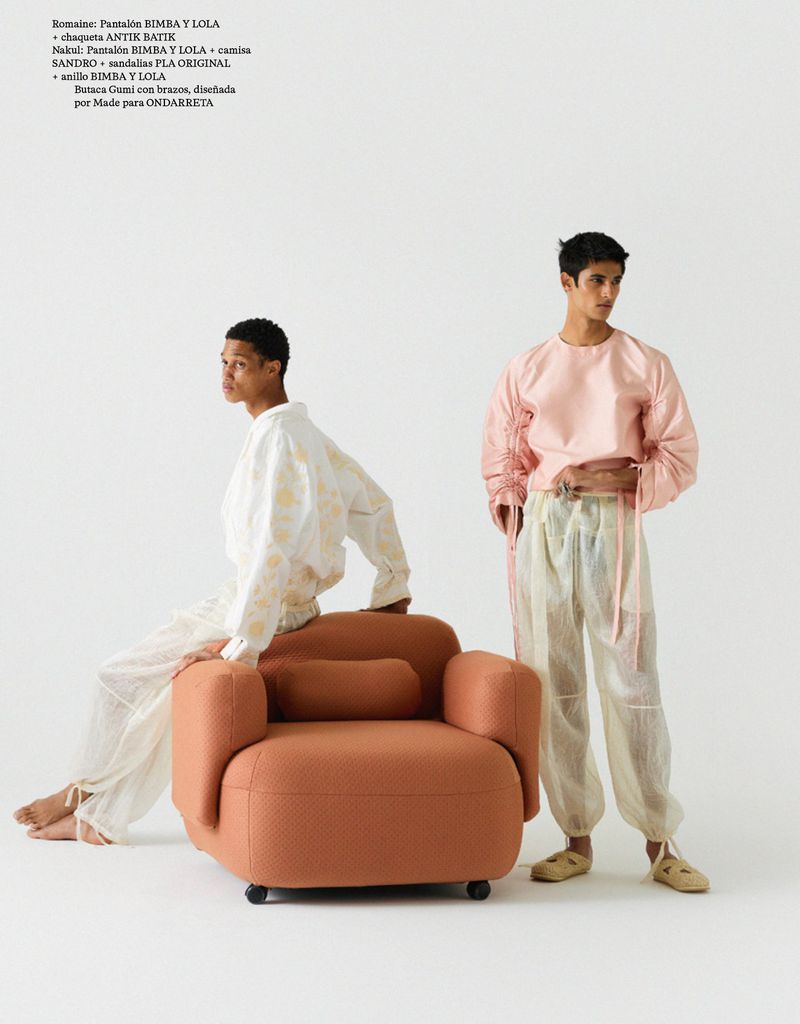 Two young adults wearing loose-fitting clothing, one seated on an orange rounded armchair in a bright, minimalist studio setting.