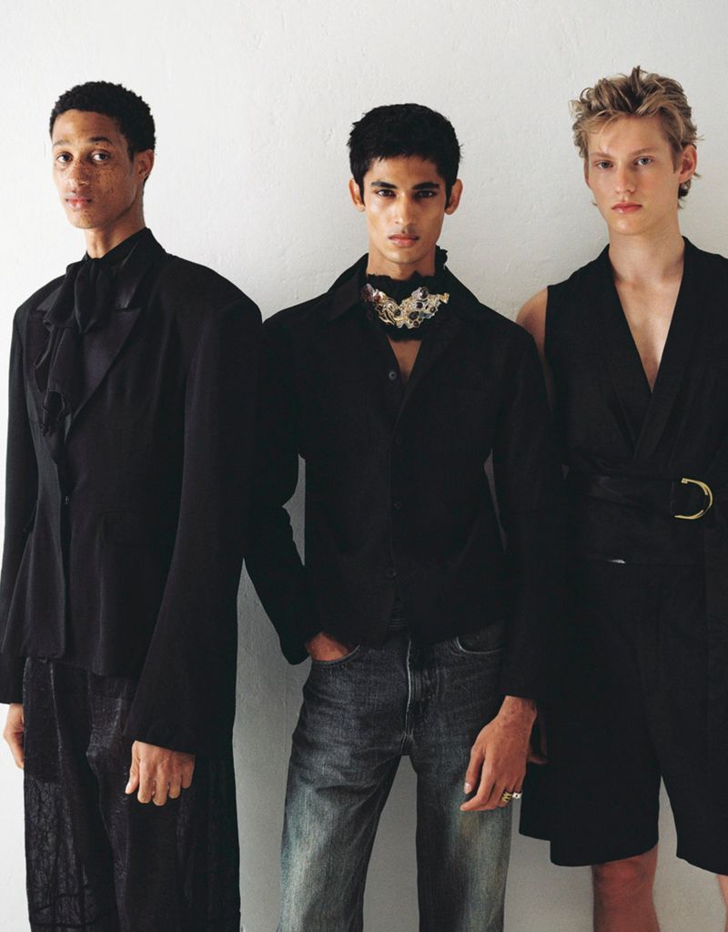 Three young male fashion models in black outfits pose against a white background in a studio setting, showcasing modern designs.