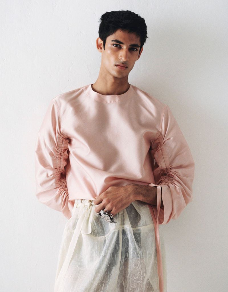 Young adult man with dark hair poses in a pink blouse with gathered sleeves and sheer ivory pants in front of a plain white wall.