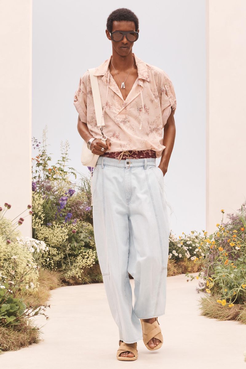 Model Craig Shimirimana walks a garden path in a pale pink short-sleeve shirt, light wash wide-leg denim, and dark sunglasses.