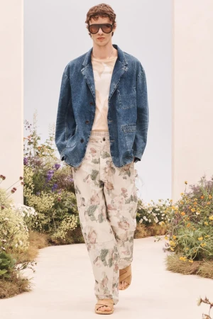 Male model walking in a blue denim jacket, beige floral print trousers, dark oversized sunglasses, and tan sandals along a garden path.