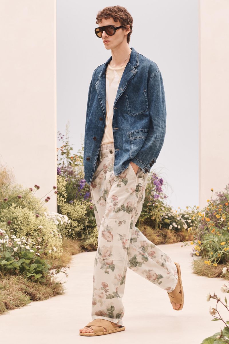 Male model walking in a blue denim jacket, floral trousers, sandals, and sunglasses on a path flanked by garden flowers.