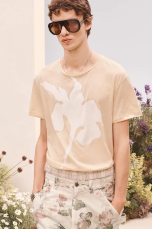 Male model wearing oversized sunglasses, a beige botanical print t-shirt, and floral trousers standing in a floral set.