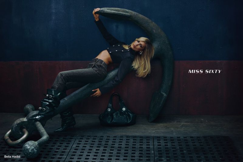 Bella Hadid in a cropped black top and dark jeans reclining against a massive ship anchor for the Miss Sixty campaign.