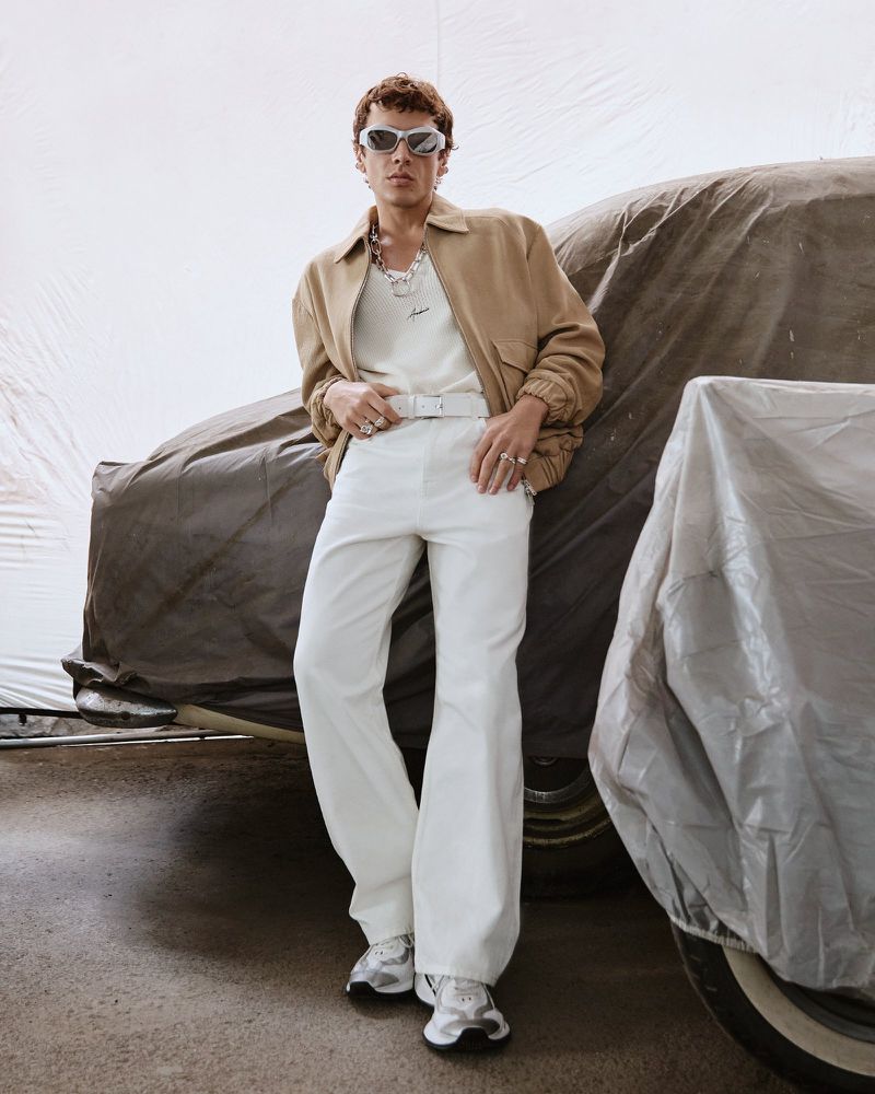 Man in neutral toned jacket, cream colored tee, and white pants poses next to a car under a tarp indoors with natural lighting.