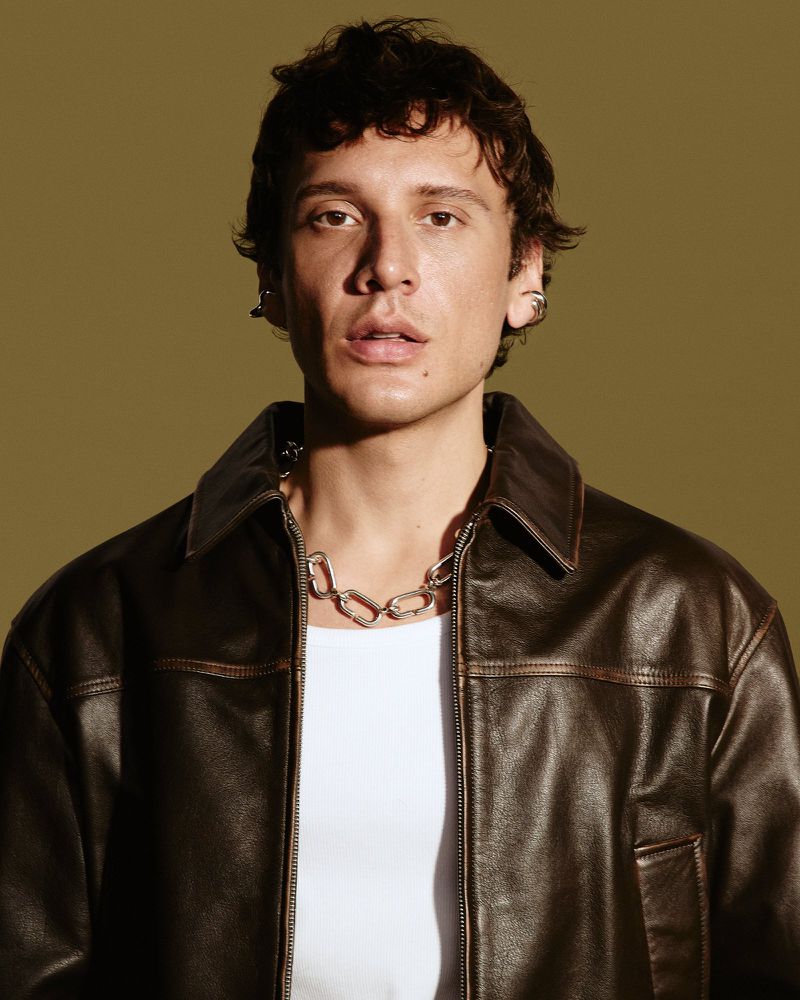 Portrait of a young adult male with dark curly hair, wearing a brown leather jacket and silver chain, against a light olive background.
