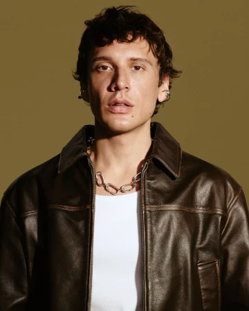 Portrait of a young adult male with dark curly hair, wearing a brown leather jacket and silver chain, against a light olive background.