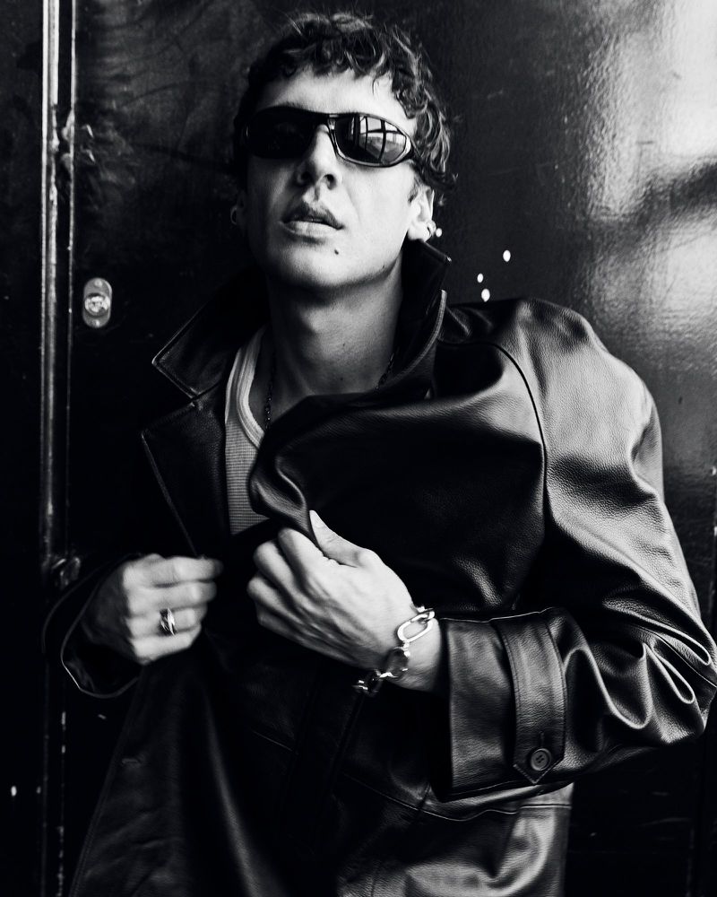Monochrome portrait of adult male wearing sunglasses and leather jacket, posing against a dark, textured wall, captured in eye-level shot.