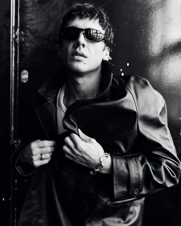 Monochrome portrait of adult male wearing sunglasses and leather jacket, posing against a dark, textured wall, captured in eye-level shot.