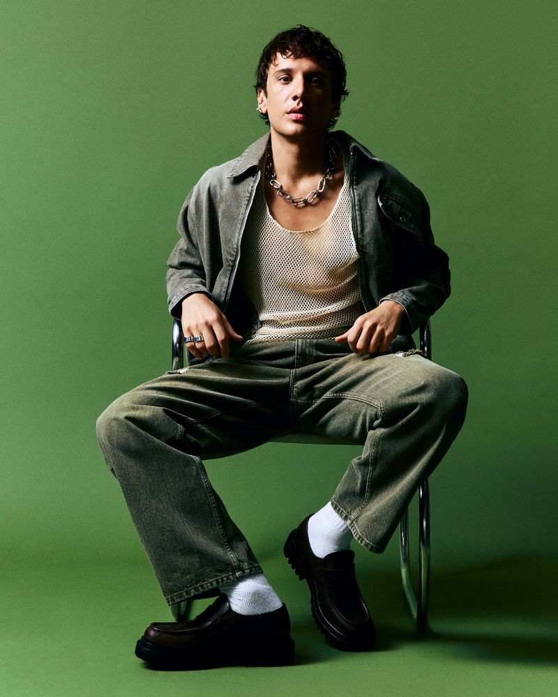 Stylish young man in jacket and jeans sitting on a chair against green backdrop, wearing a chain and chunky loafers with white socks.