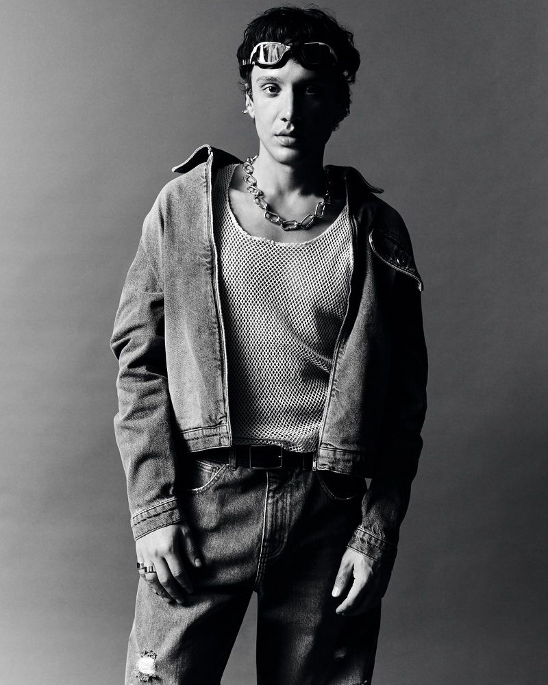 Black and white portrait of a young adult woman wearing a denim jacket, jewelry, and goggles posing against a neutral backdrop.
