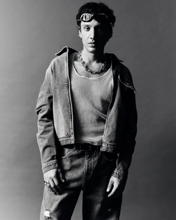 Black and white portrait of a young adult woman wearing a denim jacket, jewelry, and goggles posing against a neutral backdrop.