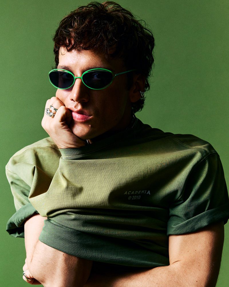 Close-up portrait of a stylish young man in a green shirt and green sunglasses, resting his chin on his hand against a green background.