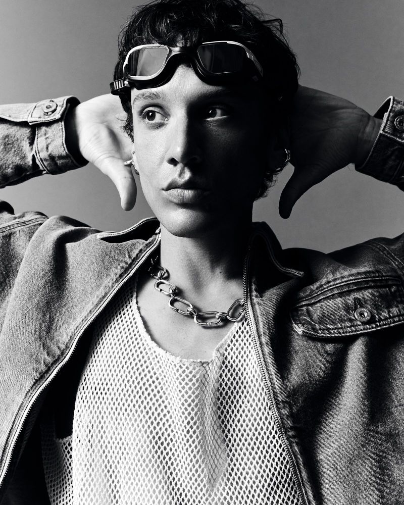Monochrome portrait of a person wearing goggles, a denim jacket, and a white mesh shirt with a silver chain necklace.