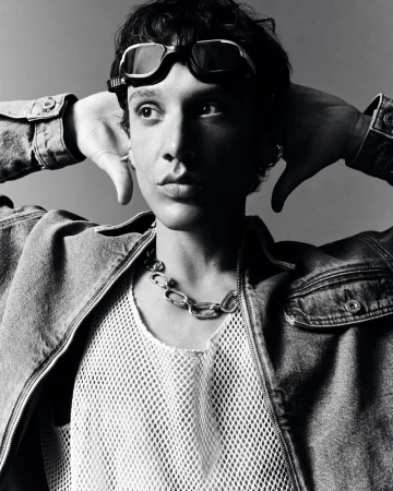Monochrome portrait of a person wearing goggles, a denim jacket, and a white mesh shirt with a silver chain necklace.
