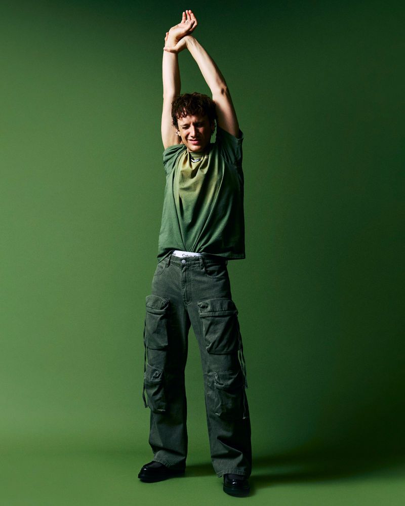 Full-body shot of a young adult man in green t-shirt and cargo pants, arms overhead, on a green background in a studio setting.